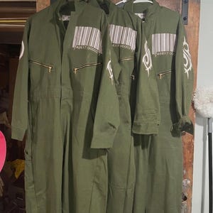 Puede incluir: Tres monos de color verde oliva colgando de perchas blancas. Cada mono tiene un código de barras impreso en el pecho y un cierre de cremallera. Un mono tiene diseños tribales blancos en la manga. Los monos se exhiben contra una puerta de madera.