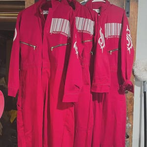 Puede incluir: Tres monos rojos colgados de perchas blancas. Cada mono tiene un código de barras y cierre de cremallera. Un mono tiene un diseño blanco en la manga. Los monos están hechos de un material duradero.