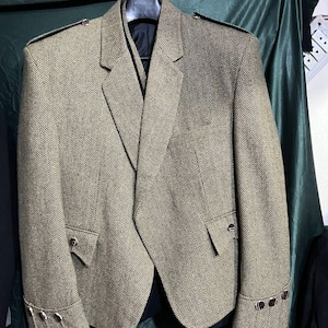Puede incluir: Chaqueta de tweed marrón y beige con diseño de espiga, solapas con muescas, dos bolsillos delanteros y botones decorativos en los puños. La chaqueta está colgada de una percha negra sobre un fondo verde oscuro.