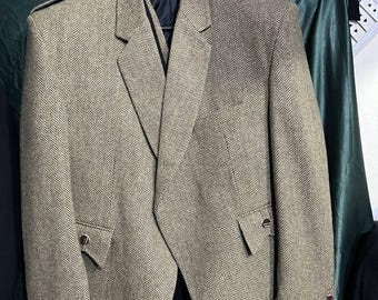 May include: A tan and brown herringbone tweed jacket with a notched lapel, two front pockets, and decorative buttons on the cuffs. The jacket is hanging on a black hanger against a dark green backdrop.