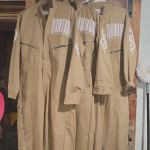 Puede incluir: Tres monos de trabajo color beige colgados de una percha. Cada mono tiene cierre de cremallera, un diseño de código de barras y un logotipo blanco en la manga. Los monos son de tela resistente y parecen nuevos.