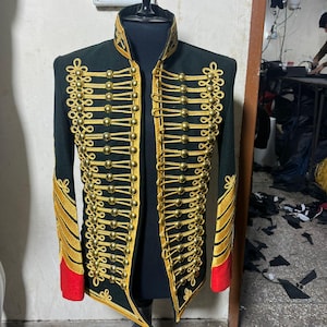 Könnte beinhalten: Dunkelgrüne Jacke im Militärstil mit goldenen Verzierungen und Knöpfen. Die Jacke hat goldfarbene Verzierungen an Kragen, Manschetten und Vorderseite, mit roten Akzenten an den Manschetten. Die Jacke wird auf einer schwarzen Schaufensterpuppe präsentiert.