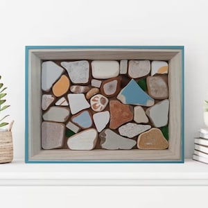 May include: A framed mosaic art piece with a blue border, filled with irregularly shaped stones in various colors like white, orange, and blue. The artwork is displayed on a white shelf, next to a potted plant and a stack of books.