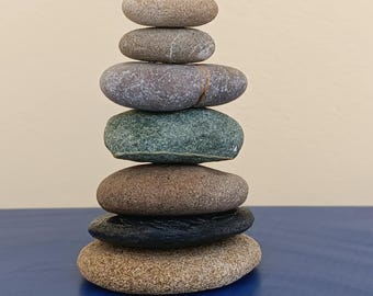 May include: A stack of seven smooth, rounded stones of varying colors, including tan, gray, green, and black. The stones are balanced on top of each other, creating a tower. The background is a solid light beige color, and the stones are sitting on a blue surface.