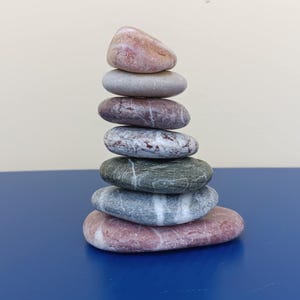 Può includere: Una pila di sette pietre lisce e arrotondate, in tonalità rosa, grigio e verde. Le pietre sono impilate l'una sull'altra, creando una torre. Lo sfondo è beige chiaro e la superficie è blu.