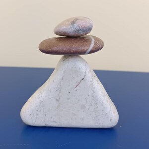 Puede incluir: Una pila equilibrada de tres piedras lisas. La piedra inferior es de color gris claro y forma triangular. La piedra del medio es marrón con una raya blanca. La piedra superior es ovalada, más pequeña, con tonos rosas y grises. Las piedras están apiladas sobre una superficie azul.