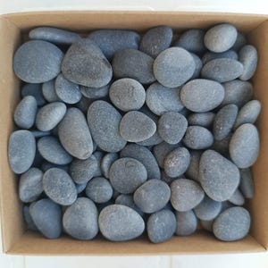 May include: A square cardboard box filled with a collection of smooth, rounded dark gray river stones. The stones vary in size and shape, creating a dense, textured layer that completely covers the bottom of the container.