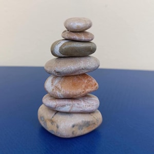 Puede incluir: Una pila de piedras lisas y redondeadas en tonos marrones y beige. Las piedras están apiladas unas sobre otras, creando una pequeña torre. El fondo es una superficie azul y una pared de color crema.