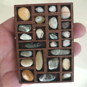 May include: A wooden display case filled with various smooth, colorful stones. The stones are in shades of beige, brown, gray, and white. The case has a dark brown finish and is divided into small rectangular compartments.