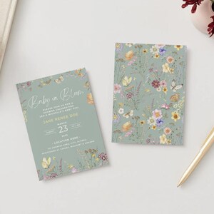 May include: Two baby shower invitations with floral and butterfly designs. One invitation reads "Baby in Bloom" with event details. The other features a floral pattern. A gold pen and a small vase with flowers are also present.
