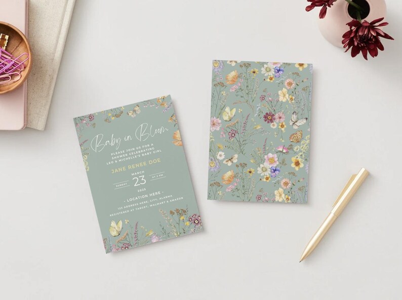 May include: Two baby shower invitations with floral and butterfly designs. One invitation reads "Baby in Bloom" with event details. The other features a floral pattern. A gold pen and a small vase with flowers are also present.