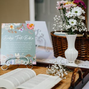May include: A table setting with a floral arrangement, a sign that reads "Baby's First Bible", and an open book. The sign is light blue with floral accents. A white vase holds colorful flowers. A woven basket is in the background.