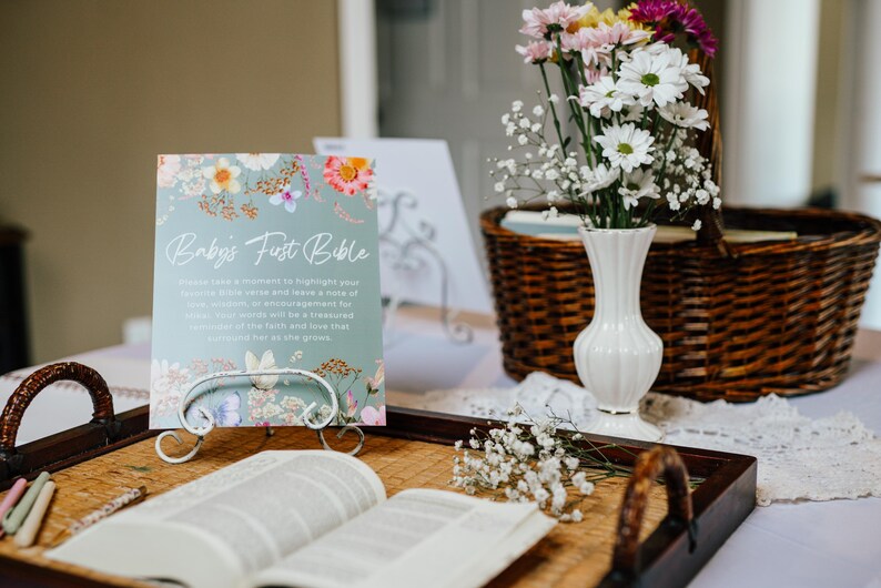 May include: A table setting with a floral arrangement, a sign that reads "Baby's First Bible", and an open book. The sign is light blue with floral accents. A white vase holds colorful flowers. A woven basket is in the background.