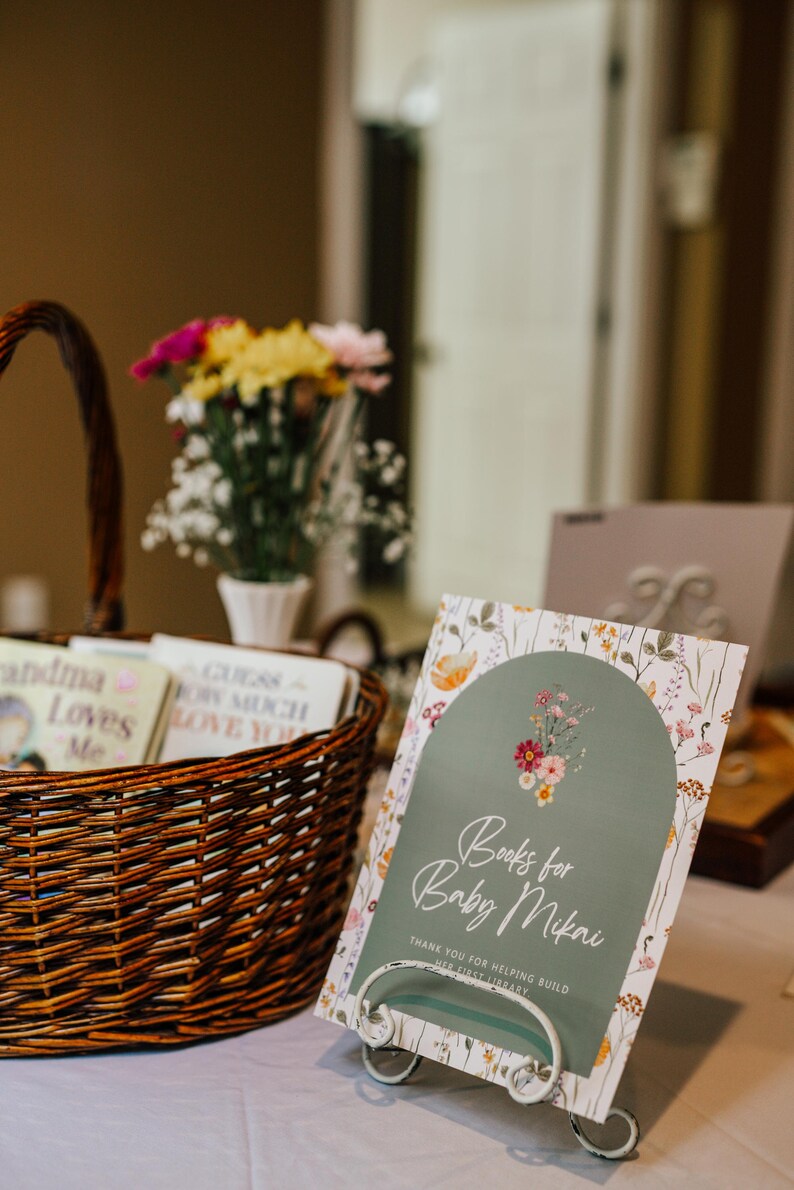 May include: A wicker basket filled with books, a sign that reads "Books for Baby Mikai", and a vase of flowers. The sign has a floral design and is on a metal stand. The books are for a baby shower.