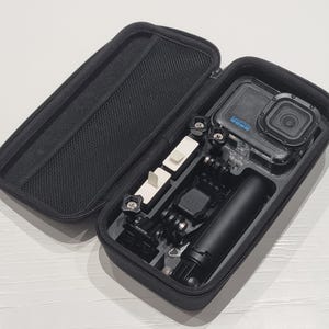 May include: Black zippered carrying case containing a GoPro camera and accessories. The case is open, displaying a camera, a handle, and various mounting components. The camera is in a clear waterproof housing. The case has a mesh pocket on the inside of the lid.