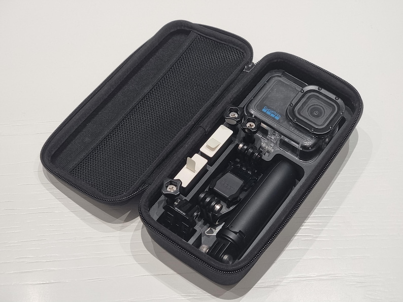 May include: Black zippered carrying case containing a GoPro camera and accessories. The case is open, displaying a camera, a handle, and various mounting components. The camera is in a clear waterproof housing. The case has a mesh pocket on the inside of the lid.