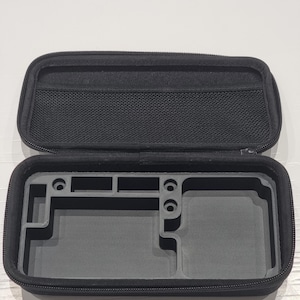May include: Open black zippered case with a mesh pocket in the lid. The interior is grey with moulded compartments, likely for a handheld gaming device. The case is designed to protect and organise the device.