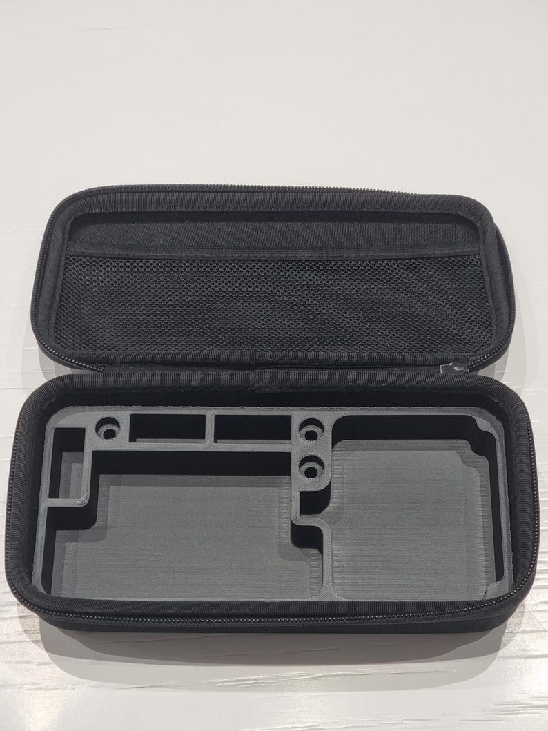 May include: Open black zippered case with a mesh pocket in the lid. The interior is grey with moulded compartments, likely for a handheld gaming device. The case is designed to protect and organise the device.