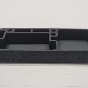 May include: A dark grey desk tidy with multiple compartments. It has a textured exterior and various sections for pens, small items, and a larger open area. The organiser is rectangular and sits on a white surface.