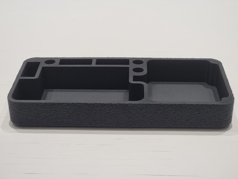 May include: A dark grey desk tidy with multiple compartments. It has a textured exterior and various sections for pens, small items, and a larger open area. The organiser is rectangular and sits on a white surface.