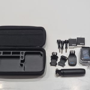 May include: A black GoPro camera kit with accessories. The kit includes a black carrying case, various mounts, a selfie stick, and two batteries. The camera is in a clear waterproof housing.