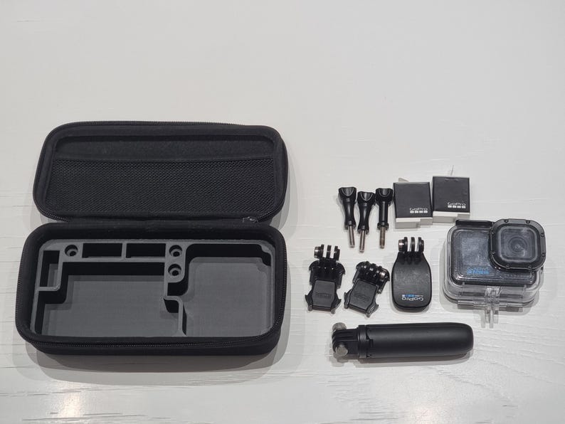 May include: A black GoPro camera kit with accessories. The kit includes a black carrying case, various mounts, a selfie stick, and two batteries. The camera is in a clear waterproof housing.