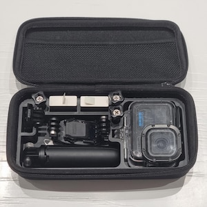 May include: Open black zippered case containing a black action camera and accessories. The case has compartments with mounting hardware, a small tripod, and a protective housing for the camera.