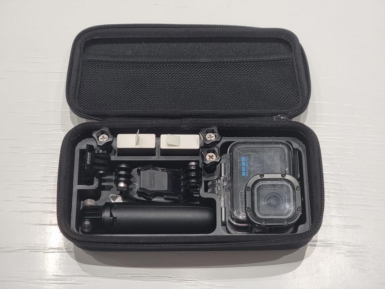 May include: Open black zippered case containing a black action camera and accessories. The case has compartments with mounting hardware, a small tripod, and a protective housing for the camera.