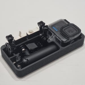 May include: Black rectangular case containing a GoPro camera and accessories. The camera is in a clear waterproof housing. Accessories include mounting hardware and a black handle. The case has compartments.