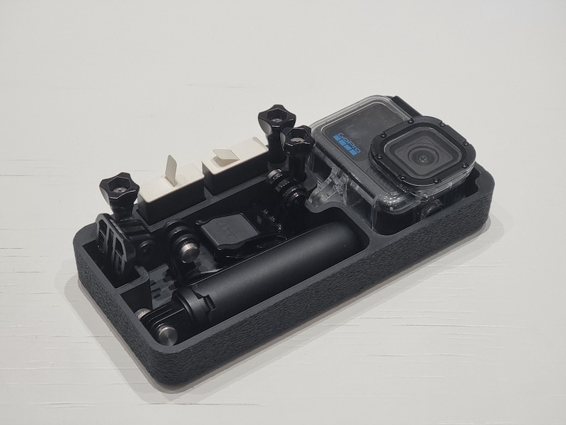 May include: Black rectangular case containing a GoPro camera and accessories. The camera is in a clear waterproof housing. Accessories include mounting hardware and a black handle. The case has compartments.