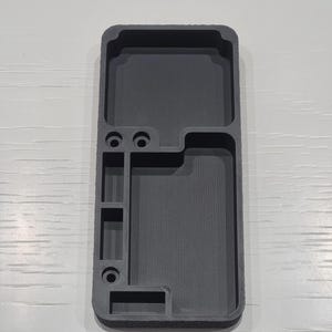 May include: A dark grey, rectangular organiser with multiple compartments and circular holes. The organiser is designed to hold various items, with a textured surface and a modern aesthetic.