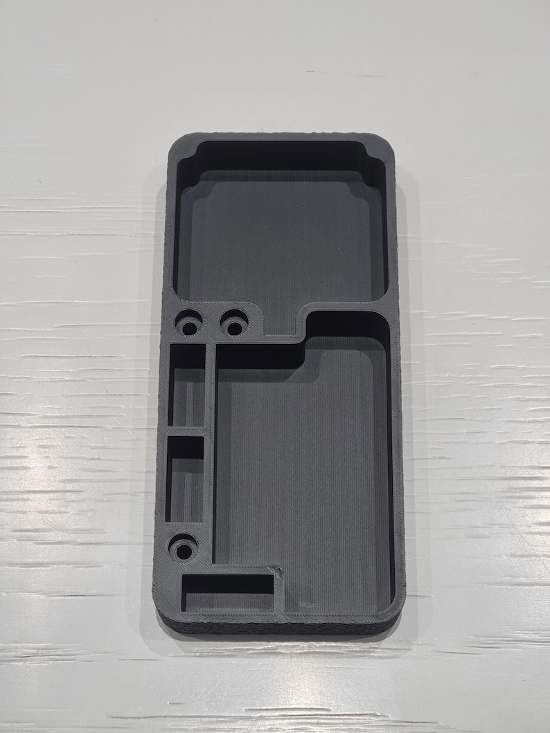 May include: A dark grey, rectangular organiser with multiple compartments and circular holes. The organiser is designed to hold various items, with a textured surface and a modern aesthetic.