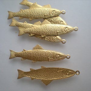 Puede incluir: Primer plano de seis colgantes de pez dorados. Cada colgante está detallado con escamas, aletas y un lazo para sujetarlo a joyas u otras manualidades. Los peces están dispuestos en un patrón ligeramente superpuesto sobre un fondo blanco.