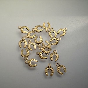 May include: A collection of small, gold-colored horseshoe charms. Each charm has a loop at the top for attaching to jewelry or other crafts. The charms are scattered on a light-colored surface.