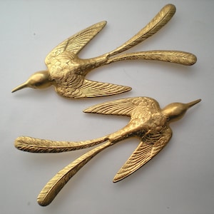 May include: Two gold-coloured metal bird figurines with detailed wings and tails. The birds are in a flying position with their wings spread out.