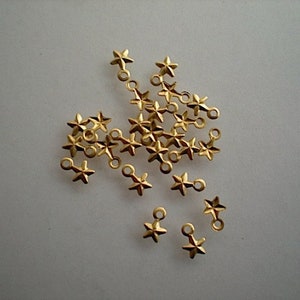 Könnte beinhalten: Eine Ansammlung kleiner, goldfarbener Sternanhänger mit einer Schlaufe zur Schmuckherstellung. Die Anhänger sind auf einem hellen Hintergrund verstreut und zeigen ihre detaillierte Sternform und das metallische Finish.