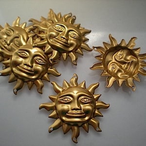May include: A set of six gold-toned metal sun charms with a face design. Each charm has a raised face with a smiling expression and a sunburst design around the face.