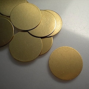 May include: A collection of small, round, brass-colored metal discs. The discs are scattered across a light-colored surface, with some overlapping. The metal has a matte finish, and the lighting creates subtle shadows.
