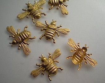 May include: Six gold-colored bee-shaped embellishments. Each bee has detailed wings, body segments, and legs. The bees are arranged on a light-colored surface, showcasing their intricate design and metallic finish.