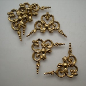 Peut inclure: Quatre équerres d'angle dorées ornées d'un motif floral et de volutes. Chaque équerre présente un élément central en forme de fleur et des bras incurvés avec de petits détails en forme de perles. Ces pièces métalliques décoratives sont probablement destinées aux meubles ou aux projets d'artisanat.