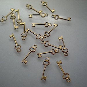 May include: A collection of gold-toned metal key charms. The keys are scattered on a white surface. The keys have a vintage style and are perfect for adding a touch of whimsy to jewelry or crafts.