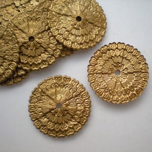 Può includere: Una collezione di abbellimenti circolari in metallo color oro. Ogni pezzo presenta un design dettagliato, simile a un fiore, che si irradia da un foro centrale. Gli abbellimenti sono disposti su una superficie bianca, mostrando la loro intricata consistenza e forma.