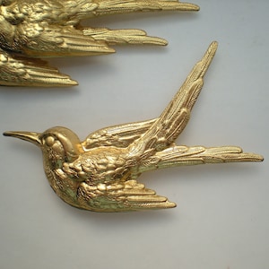 Puede incluir: Dos figuritas de pájaros de metal dorado con alas detalladas y cuerpos texturizados. Los pájaros están en posición de vuelo con las alas extendidas.