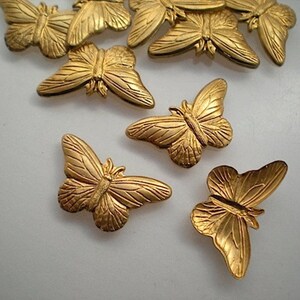 May include: A collection of small, gold-colored butterfly embellishments. The butterflies have detailed wing patterns and are arranged on a white surface. The wings are slightly spread, and the overall design is ornate.