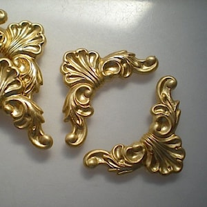 May include: Gold-colored, ornate decorative corner pieces with a detailed, swirling leaf design. The pieces are likely for furniture or decorative applications, with a classic, vintage aesthetic. The pieces are made of a solid material.
