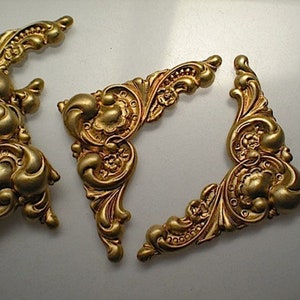 Puede incluir: Tres piezas de esquina de metal de color dorado con intrincados diseños florales. Las piezas son ornamentadas y tienen un aspecto vintage.