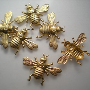 May include: Six gold-colored metal bee charms with detailed wings and bodies. The bees are arranged in a cluster on a white background.