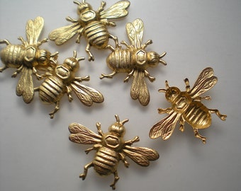 May include: Six gold-colored metal bee charms with detailed wings and bodies. The bees are arranged in a cluster on a white background.