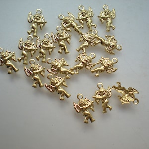 May include: A collection of 18 gold-toned metal charms featuring cherubs with wings, holding arrows. The charms are scattered on a white surface.