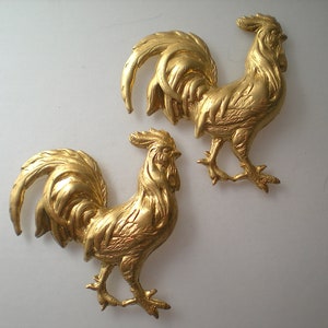 May include: Two gold-colored rooster figurines with detailed feathers and a standing pose.